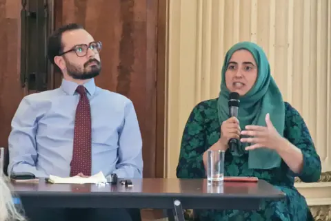 LDRS A man in a shirt and tie sitting next to a woman, wearing a headdress and talking into a microphone. 