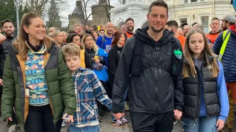 Craig Maxwell with his wife and two kids. There is a crowd of people behind him.