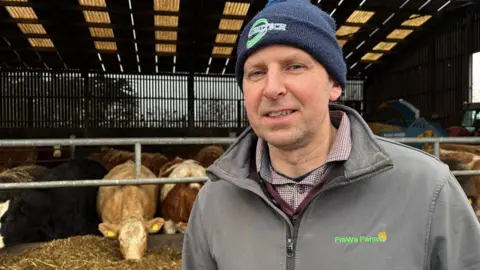 Fergal Watson, a man with short hair, looks to the camera on his farm in Cloughey, County Down. He is wearing a blue beanie hat with a green logo, a grey zipped jacket and a red checked shirt. He is standing in front of a large cattle shed. Cattle are eating silage from a feeding trough.