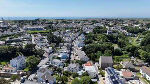 A picture of Alderney taken from above. There is a number of houses pictured with trees surrounding it.