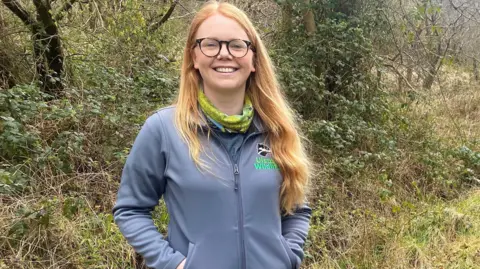 BBC NI Katy is standing in a wooded area. She wears glasses and has long auburn hair. She's wearing an Ulster Wildlife jacket and a scarf.