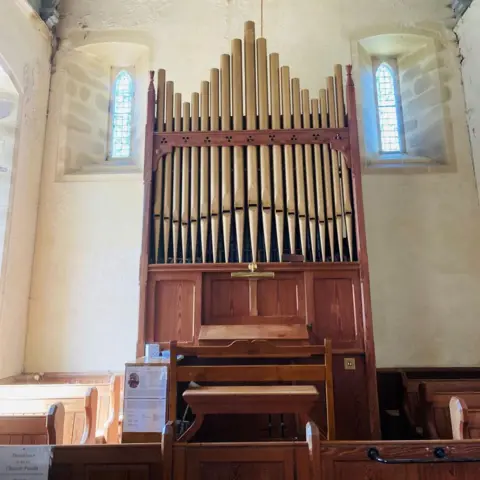 Large brown musical organ inside church building, with light streaming through narrow windows.