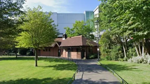 A path, flanked by patches of neat grass and trees, leads to a small building housing a public toilet. A large, more modern building can be seen rising in the background. 
