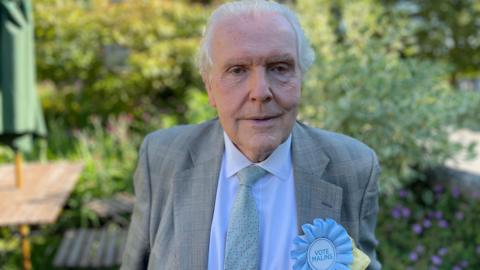 Julian Malins is wearing a grey suit with tie and a light blue rosette, standing in a garden