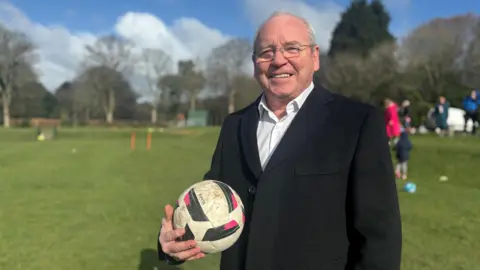 A man in his 60s, clean-shaven, wearing glasses and with thinning grey hair, stands on a grass field in a park. He is wearing a black blazer over a white shirt and holds a football in his right hand as he smiles for the camera.