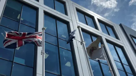 A series of three flags fluttering outside the glass fronted Slough Borough Council office building, Observatory House. One of the flags has the council logo on it and in the top right corner you can see a few clouds in what is a very bright sky