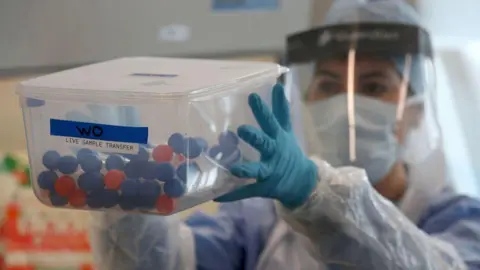 An NHS worker is holding a box of coronavirus tests. She is wearing a visor, gloves and an apron. You cannot see her face because she is also wearing a mask.
