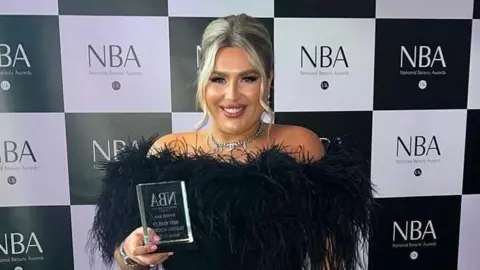 Charlotte Bates with blond hair worn up wearing an evening black velvet dress with black feathers on the collar. She is holding up a glass trophy she won at te National Beauty Awards.