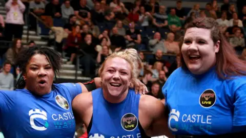 Giants Live Three women in blue T shirts. The first woman has black curly hair, the second woman has tied back blonde hair and the third woman has red and brown hair. They are all smiling at the camera