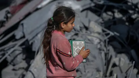 EPA A young girl holds a copy of the Quran at the site of the destroyed Al Bokhari mosque following an Israeli airstrike in Deir Al Balah, southern Gaza Strip