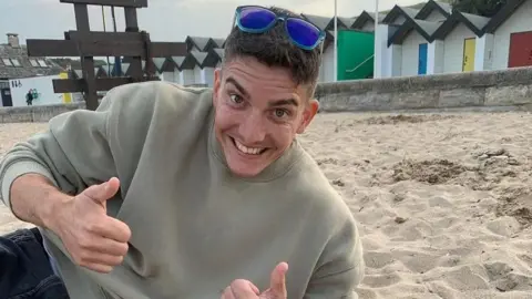 Family handout Adam Harrison lies down on a sandy beach with multiple beach huts in the background. He has a broad smile and is doing a two thumbs up gesture. He has blue sunglasses propped on his head and is wearing jeans and a light green jumper.