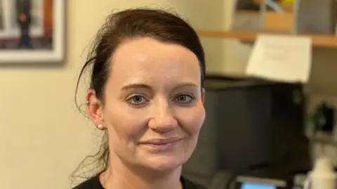 Gill Dummigan/BBC A woman with tied-back brown hair smiles at the camera while sat at an office desk