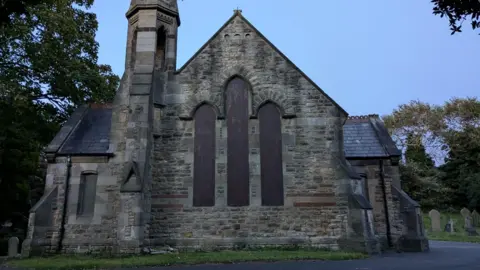 Ryton Chapel. The stone building's spire can be seen as well as three of its tall windows have been boarded up. Gravestones lie on either side of the building.