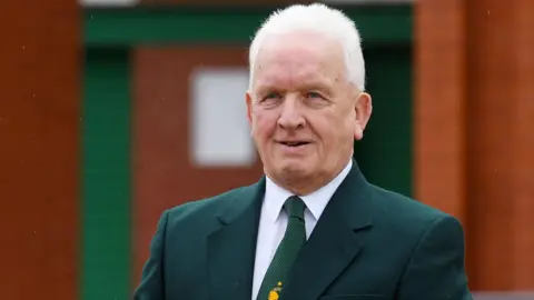 SNS A man with white hair and a green suit jacket and tie gives a small smile to the camera