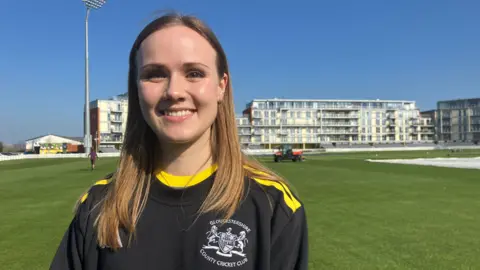 A women is smiling at the camera. She is wearing a Gloucestershire County Cricket Club branded t-shirt which is black with yellow stripes across the shoulders and around the neck. She has mid-length, light brown hair. She is stood on a cricket ground. 