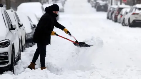A man rugged up in heavy winter clothing uses a tool to clear snow packed into a shovel he is using to clear part of a road near a line of parked cars, all of which have snow over their roofs and windscreens.