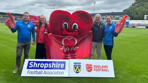 Four people are standing on a football pitch. In the middle is a person dressed as a giant red heart. There is a flag that reads "British Heart Foundation". Two of the people have red foam fingers on their hands.