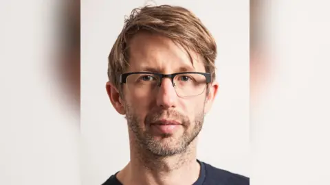 Paul Arnold Paul Arnold has light brown hair and stubble. He is wearing glasses and a black T-shirt.