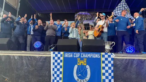Philomena Begley who is wearing a denim jacket and jeans sings on stage flanked by players and staff from Dungannon Swifts with a Swifts blue and white flag draped over the front of the stge