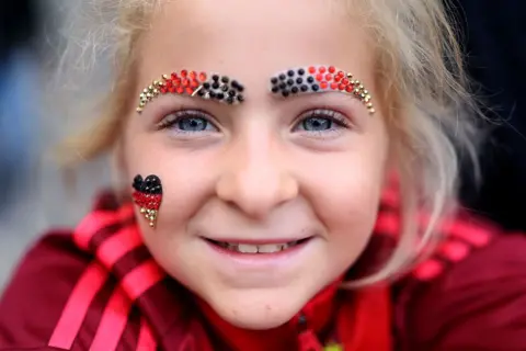 Getty Images A young girl with blonde hair and blue eyes smiles to the camera in a close up shot. She is wearing sparkly eyebrows made out of beads in the colours of the German flag and a little heart on her right cheek