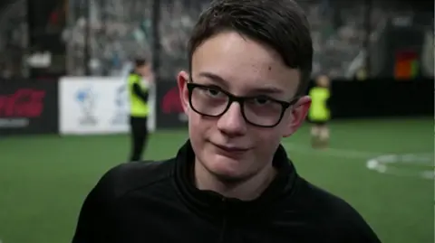Buddy stands on an indoor football pitch. He is wearing a black sports-style top with a zip at the collar. The background features artificial turf, along with several people engaged in football activity. Some of them are dressed in bright training bibs, and one person appears to be standing near the centre of the pitch. Advertising boards and a goalpost are visible in the distance, giving the scene the atmosphere of an indoor training facility or sports centre.