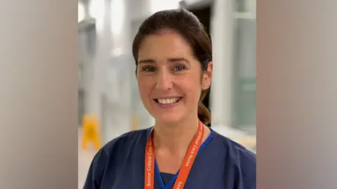 A woman who brown hair in a ponytail smiles at the camera, wearing a blue overall with an orange lanyard, standing in the corridor of a hospital 