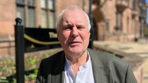 George Duggins has short white hair. He wears a grey suit jacket and a white shirt.