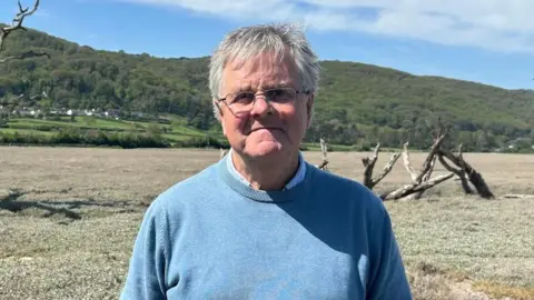 A man in a blue jumper is looking ahead. He has short grey hair and glasses on. Behind is a forested area, and closer, there is marsh land.
