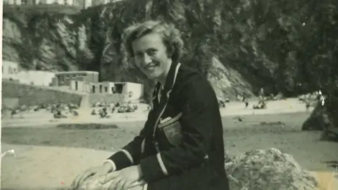 Mary on a beach, wearing a smart blazer and smiling at the camera. 