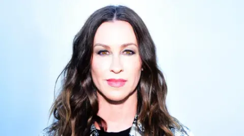 Alanis Morissette stands against a light blue background. She has long wavy dark hair and is wearing a black and white collared shirt. 
