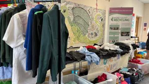 A view of school uniform items on a rail to the left, and on the table, a range of different coloured clothing on a table. Underneath the table are boxes filled with more items. 