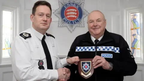 Essex Police Chief Constable Ben-Julian Harrington, in a police issue white shirt and black tie, hands Supt Rayner, dressed in the Specials uniform, a wooden plaque with the police badge on it. They are in a white room, with the Essex Police badge on the wall behind them.