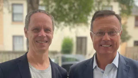 Frankie Golding/BBC Sebastian Warrack and Robin Smith are both wearing blue jackets and smiling as they stand in a street outside Jersey Opera House with a pale cream building behind them.