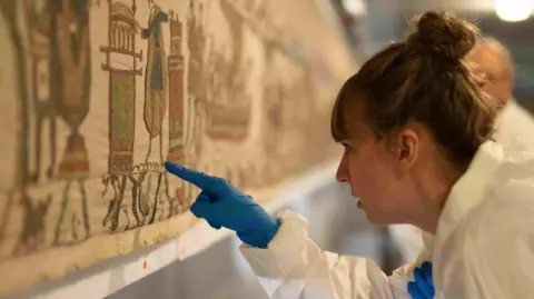A woman in a sterile suit and gloves inspects the artwork with a pointed finger.