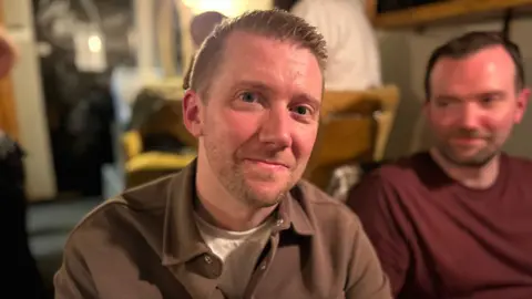 A man with short spiky brown hair looks directly at the camera. He has a slight smile on his face and is wearing a brown shirt and white t-shirt underneath. To his right is another man looking at him with a maroon coloured top on and short brown hair. 