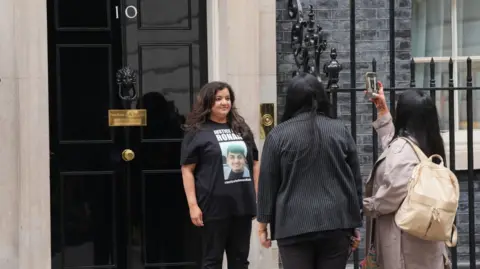 PA Media Ms Kanda is stood outside Downing Street having her photo taken by two women. She is wearing black T-shirt with her son's face on the front.