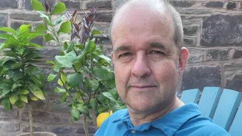 Nick Price A man sits outdoors on a chair beside potted plants and a brick wall.
