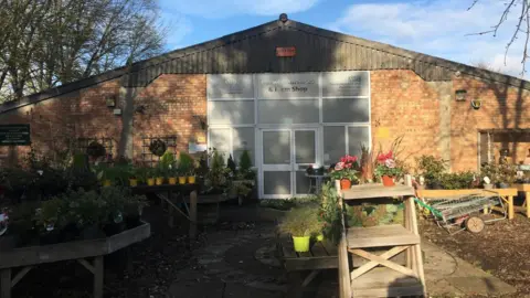 A large triangular brick building with a tinted glass door and windows that say "Darwin Nurseries & Farm Shop". In front of the building are several stalls with potted plants on them. 