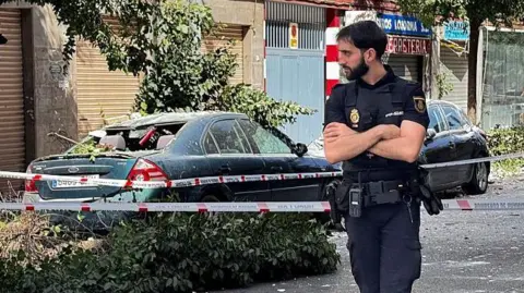 A policeman with his arms crossed stands on a street, and a very damaged car is behind him.