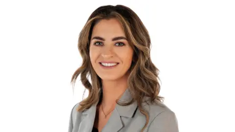 A woman with long brown curly hair, smiling, wearing a grey blazer pictured against a white wall