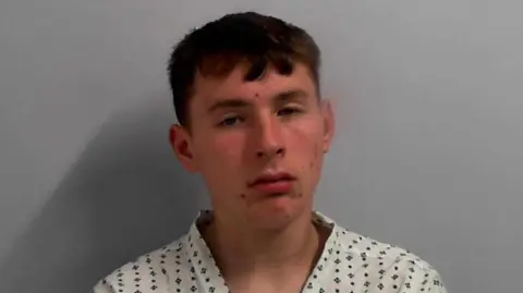 A mugshot of a young man wearing a white shirt with a blue pattern on it. 