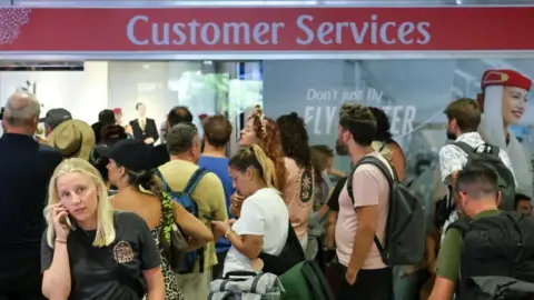 Travellers at Dubai airport queue while looking stressed, underneath a sign that reads 'customer services'