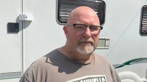 Alexander Stevanovic/BBC Barry is wearing black-framed glasses and a brown T-shirt. He is standing in the sunshine with a white caravan behind him.