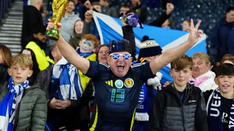 PA Media A fan wearing a Scotland football jersey holds up a replica World Cup trophy and makes a victory gesture, surrounded by other supporters with scarves and flags in a stadium.