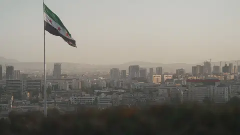 A cityscape surrounded by mountains in the distance is captured in a red dusk like light. A single flag pole has a large flag catching the wind. It has a green stripe at the top, a white stripe bearing three red stars, and a black stripe at the bottom.