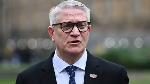 Getty Images Andrew Rosindell is pictured wearing glasses and a dark suit and tie speaking outdoors, with trees and railings blurred in the background.