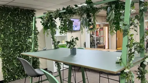 A high table with four green and grey high chairs sat around it. Over the table is a green frame, with a bower made of flowers and leaves wrapped around it. On the far wall is a carpet of leaves and ivy. 