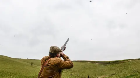 Getty Images A man wearing a brown jacket and flat cap aims a gun into the air where there are pheasants flying.  