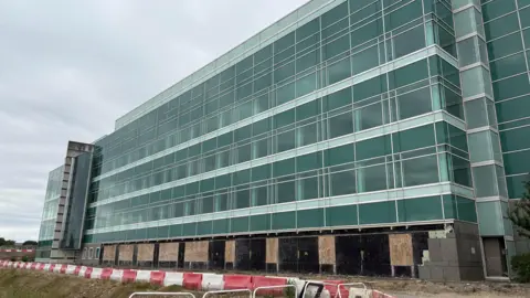 New National Biosecurity Centre in Harlow aims to fight pandemics 6 Ben Schofield/BBC Modern multi-story building with glass windows, the ground floor us under construction with boarded-up sections.