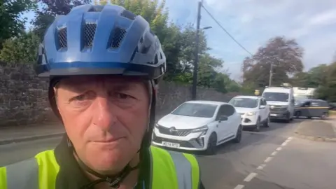Frank Biederman Frank Biederman in a hi-vis jacket and blue cycling helmet with queuing cars behind him